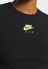 Černé bavlněné tričko s kulatým výstřihem a krátkými rukávy. Na hrudi má žluté logo Nike a text "AIR". Hladká textura.