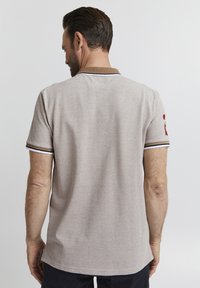 Camisa polo beige con un cuello marrón y rayas blancas, con mangas cortas y un detalle del logo rojo en la manga izquierda.