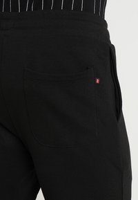 Black cotton joggers featuring a back pocket with a small red logo accent. The fabric has a smooth texture and elastic waistband.