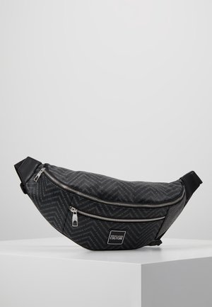 Sort Versace Jeans Couture crossbody taske med diagonalt logoprint og to sølv lynlåse rum på en hvid piedestal.