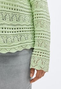 Ανοιχτό πράσινο πλεκτό πουλόβερ με σχέδια δαντέλας, κυματιστό τελείωμα και μακριά μανίκια. Φοράται πάνω από γκρι ύφασμα.