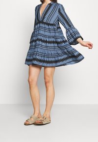 Robe noire et bleue à motifs avec un décolleté en V profond, des manches évasées et une jupe à volants. Associée à des sandales beiges à boucles.