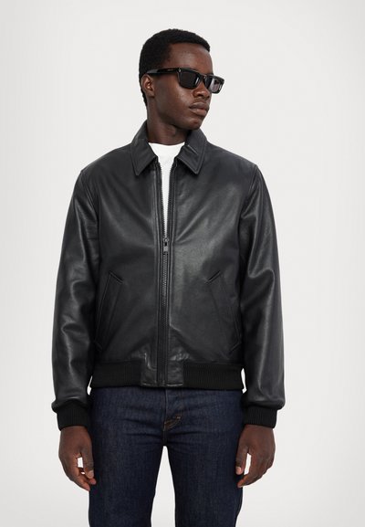 Chaqueta bomber de cuero negro con cuello tipo solapa, cremallera frontal y puños de canalé. Llevada sobre una camisa blanca, combinada con jeans de mezclilla oscura.