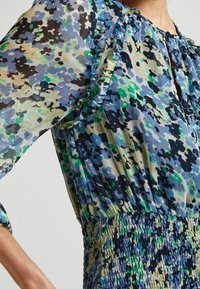 Zbliżenie sukienki z abstrakcyjnym kwiatowym wzorem w odcieniach niebieskiego, zielonego, czarnego i beżowego, z falbaniastymi krawędziami rękawów oraz marszczonym detalem w talii.