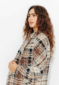 Veste à carreaux marron et blanc en tissu texturé, dotée d'un col et d'un bouton, avec des accents noirs et une coupe décontractée.