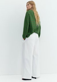 Donna con capelli biondi lunghi che indossa un maglione a righe verdi, pantaloni bianchi larghi e tacchi neri aperti, in piedi contro un muro bianco.