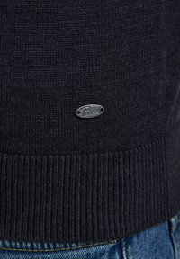 Ciemnoniebieski sweter z dzianiny z ribbanym dołem, z małą srebrną metką "Petrol" umiejscowioną blisko dolnej krawędzi. Widoczne dżinsy poniżej.