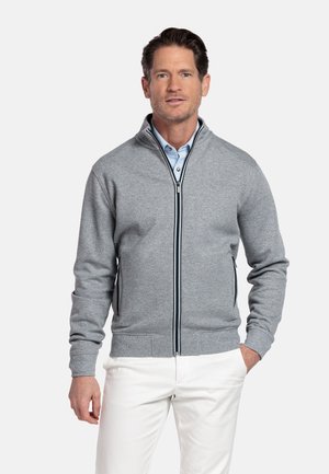 Man met lichtblauw overhemd met knopen, grijze jas met ritssluiting en witte broek, staand met één hand in de zak tegen een witte achtergrond.