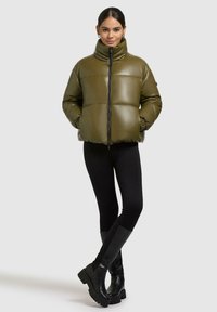 Kvinde iført en olivengrøn dunjakke med lynlås, sorte leggings og sorte knæhøje chunky støvler, stående med hænderne i lommerne.