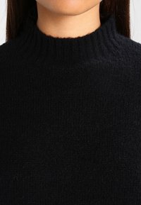 Vrouw draagt een zwarte, geribde gebreide trui met een hoge, licht golvende halslijn, weergegeven van nek tot bovenborst.