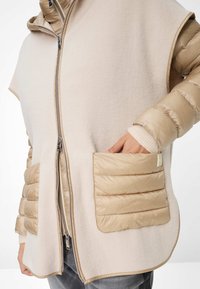 Beige wollen blend buitenkleding met gevoerde beige schouders en zakken, voorzien van een volledige ritssluiting en geribbelde manchetten. Twee grote opgestikte zakken zichtbaar.