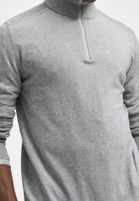 Grå stickad tröja med hög krage, halv dragkedja och ribbade detaljer vid ärmsluten. Mjuk textur och figursydd design.