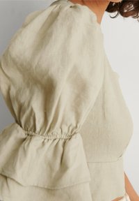 Haut beige à manches bouffantes avec fronces élastiquées sur le haut du bras et au poignet, porté par une personne vue de profil sur un fond uni.