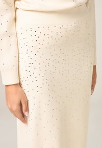 Gonna in maglia color crema decorata con brillantini sparsi, caratterizzata da una texture liscia e una silhouette dritta.