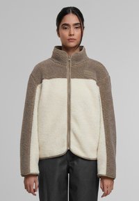 Chaqueta de forro polar ligera; presenta un cuerpo color crema con acentos marrones, cuello alto, cremallera completa y una superficie texturizada.