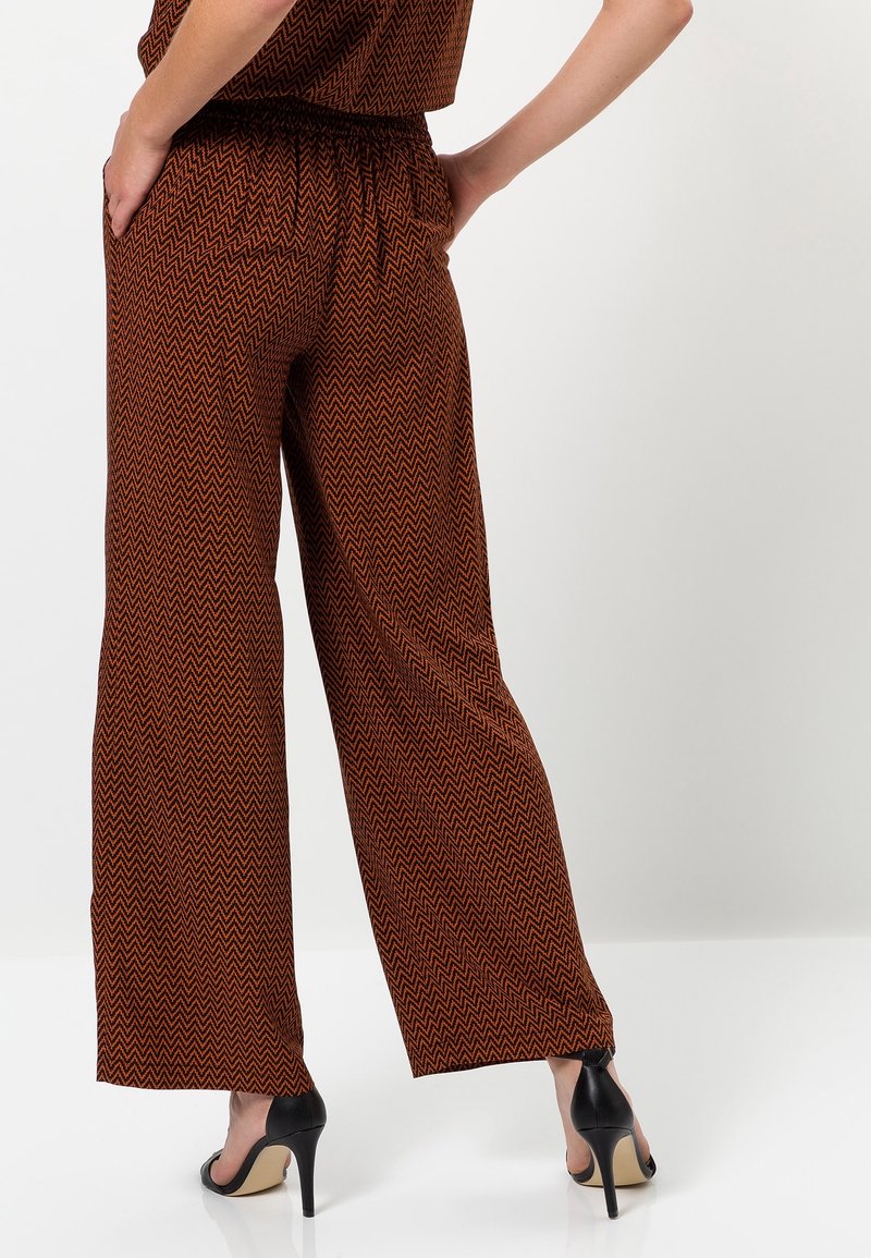 Personne portant un pantalon large marron à motif zigzag et des chaussures à talons noires, debout les mains dans les poches, fond blanc.
