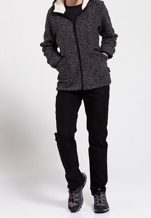 Person iført en mørkegrå fleecetrøje med lynlås og lommer, sorte bukser og grå vandresko, stående med hænderne i lommerne.