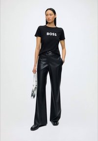 Kvinde står iført en sort "BOSS" t-shirt, sorte læderbukser med vide ben, sorte sko og bærer en lille sølv håndtaske.