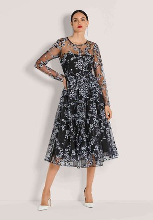 Frau trägt ein schwarzes Midikleid mit durchsichtigen, floral bestickten langen Ärmeln und weißen spitzen High Heels, posiert vor einem schlichten Hintergrund.