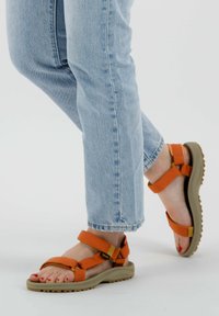 Sandalias naranjas con tiras ajustables, suela de goma y una plantilla texturizada. Combinadas con jeans de color azul claro.
