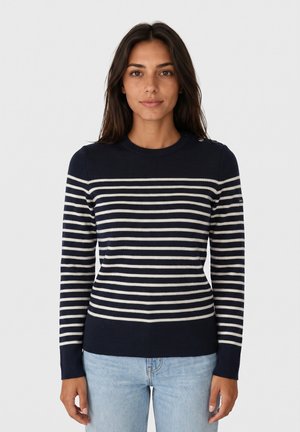 Donna con capelli scuri che indossa un maglione blu navy con strisce orizzontali bianche e jeans azzurri, in piedi davanti a uno sfondo chiaro e uniforme.
