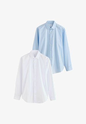 Mann trägt hellblaues Hemd, dunkelgraue Hose und schwarze Anzugschuhe, steht vor einfachem weißem Hintergrund.