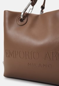 Brązowa torba na ramię z skóry o fakturowanej powierzchni, z wytłoczonym logo, srebrnymi elementami i okrągłym detalem na uchwycie.