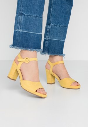 Żółte zamszowe sandały na wysokim obcasie z odkrytymi palcami i regulowanym paskiem na kostkę. Posiadają okrągłą klamrę i masywny obcas.