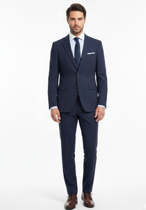 Hombre de pie vistiendo un traje azul marino hecho a medida con camisa blanca, corbata azul marino con lunares, pañuelo blanco en el bolsillo y zapatos de vestir marrones sobre fondo blanco.