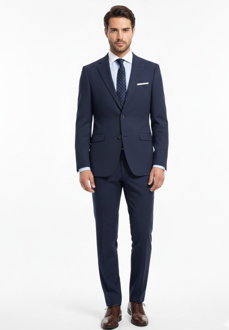 Hombre de pie vistiendo un traje azul marino hecho a medida con camisa blanca, corbata azul marino con lunares, pañuelo blanco en el bolsillo y zapatos de vestir marrones sobre fondo blanco.