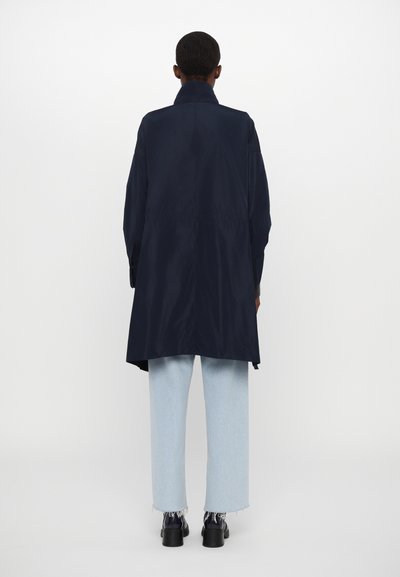 Tumesinine trench coat, millel on pikk, sujuv siluett ja krae, koos helesiniste sirgete säärtega teksade ja mustade kingadega.