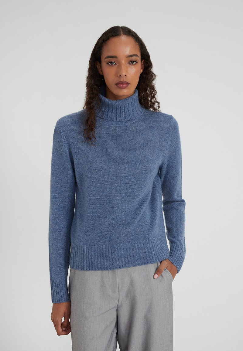 Maglione turtleneck blu in morbido tessuto a maglia con polsini e orlo a coste, indossato sopra pantaloni grigi su misura, caratterizzato da una vestibilità rilassata.