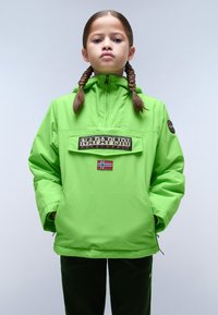 Bambino con capelli intrecciati che indossa una vivace giacca Napapijri verde brillante con patch della bandiera norvegese, mani in tasca, in piedi contro uno sfondo neutro.