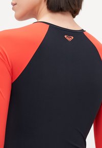 Piros ujjú, fekete testű rash guard; a nyakrészen egy kis logó található. Simaságot és rugalmasságot biztosító anyagból készült. Kényelemre és mozgékonyságra tervezték.