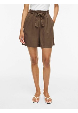 Žena nosiaca hnedé kraťasy s vysokým pásom so zviazaným látkovým opaskom, bočné vrecká, biele sandále a biele tričko, stojaca na jednofarebnom pozadí.