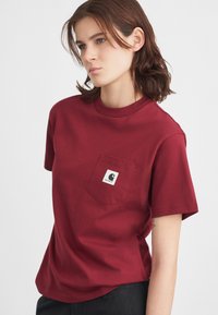 T-shirt in cotone bordeaux con collo rotondo, maniche corte e taschino sul petto con una patch logo a contrasto. Tessuto liscio, vestibilità comoda.
