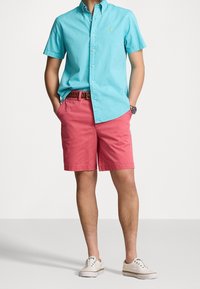 Ljusblå skjorta med knappar, rosa shorts, brun bälte och vita sneakers. Korta ärmar, avslappnad passform och enstaka färger utan mönster.