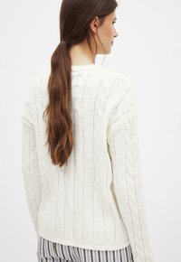 Pull en maille torsadée blanc avec texture côtelée, doté d'un col rond et de manches longues, porté sur un pantalon rayé.