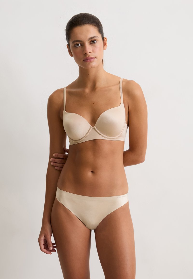 Frau mit dunklen Haaren in einem beigen BH und passender Unterwäsche steht mit einem Arm hinter ihrem Rücken vor einem schlichten hellen Hintergrund.