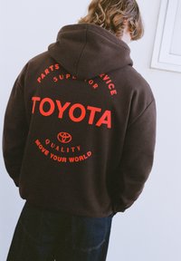 Hnedá mikina s kapucňou s červeným nápisom. Text obsahuje "TOYOTA," "KVALITA" a "POSÚVAJ SVOJ SVET." Má sťahovaciu šnúrku na kapucni a voľný strih.