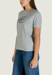 Szara bawełniana koszulka z krótkim rękawem, z czarnym haftowanym logo „CK” na piersi. W połączeniu z niebieskimi dżinsami.