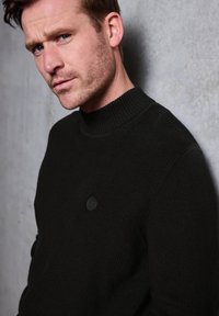 Pull en maille noir avec un col roulé haut, arborant un badge logo discret sur le devant. La texture est douce et ajustée.