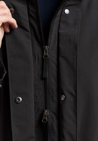 Svart zip-up jacka med en robust dragkedja, tryckknappar och en slät yta. Nära vy av framsidan och det inre fodret.