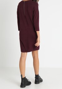 Vestido de suéter burdeos con mangas tres cuartos, escote redondo y detalle de cremallera en la parte posterior, combinado con botines negros. 