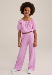 Svetlo roza, teksturiran outfit, ki vključuje krajšan top s pufastimi rokavi in široke hlače. Stilizirano z belimi supergami.