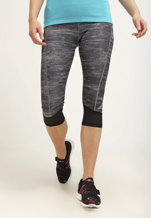 Femme portant des leggings capri adidas gris et noirs et des chaussures de sport noires marchant devant un fond clair uni.