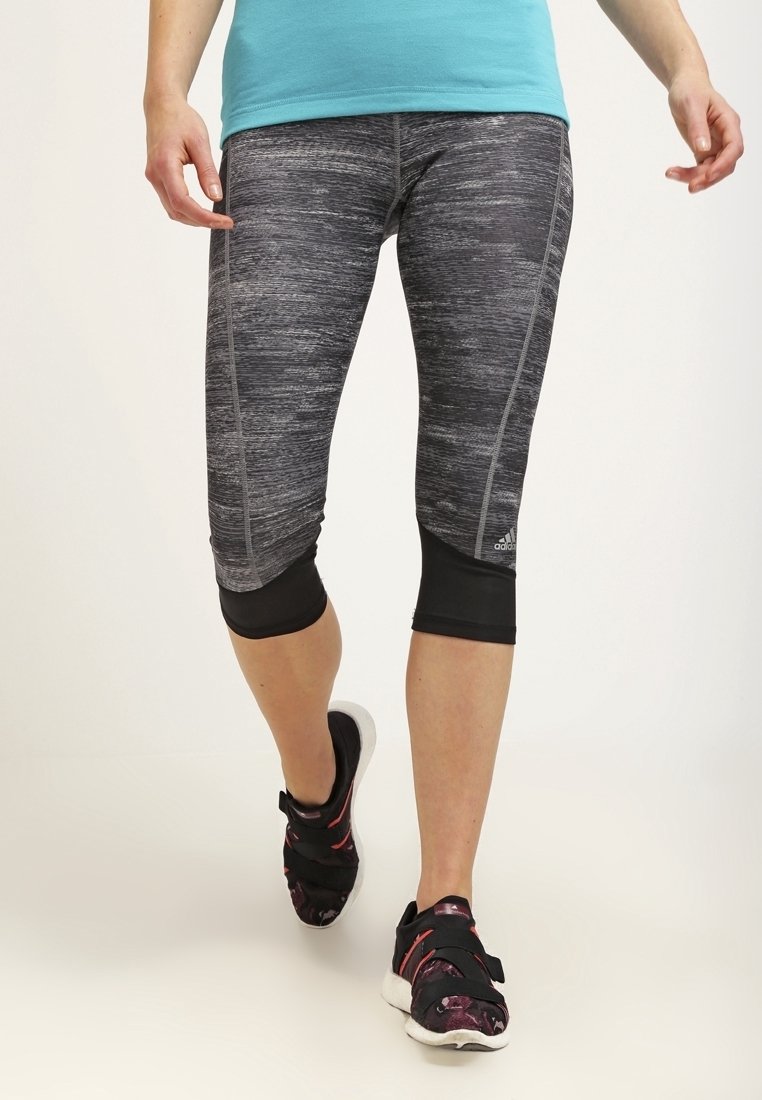 Femme portant des leggings capri adidas gris et noirs et des chaussures de sport noires marchant devant un fond clair uni.