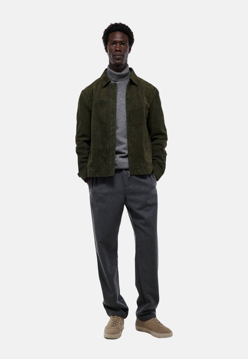 Veste en daim verte, pull à col roulé gris, pantalon en laine gris, baskets beiges. Un homme se tient droit avec les mains dans les poches de la veste, sur un fond neutre.
