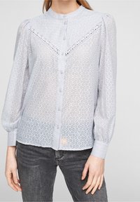 Blusa azzurra chiara realizzata in un tessuto leggero e decorato, con chiusura a bottoni frontale, maniche lunghe a sbuffo e dettagli in pizzo decorativo sulle spalle.