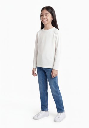 Mosolygó gyermek hosszú sötét hajjal, fehér hosszú ujjú inget, kék farmert és fehér tornacipőt visel, fehér háttér előtt áll.